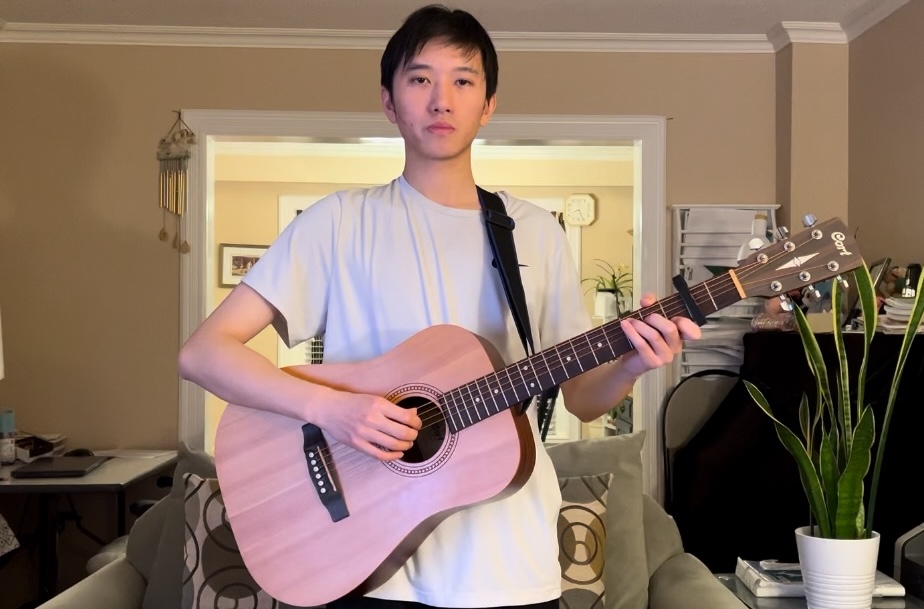 Person wearing white t-shirt playing a wooden acoustic guitar standing